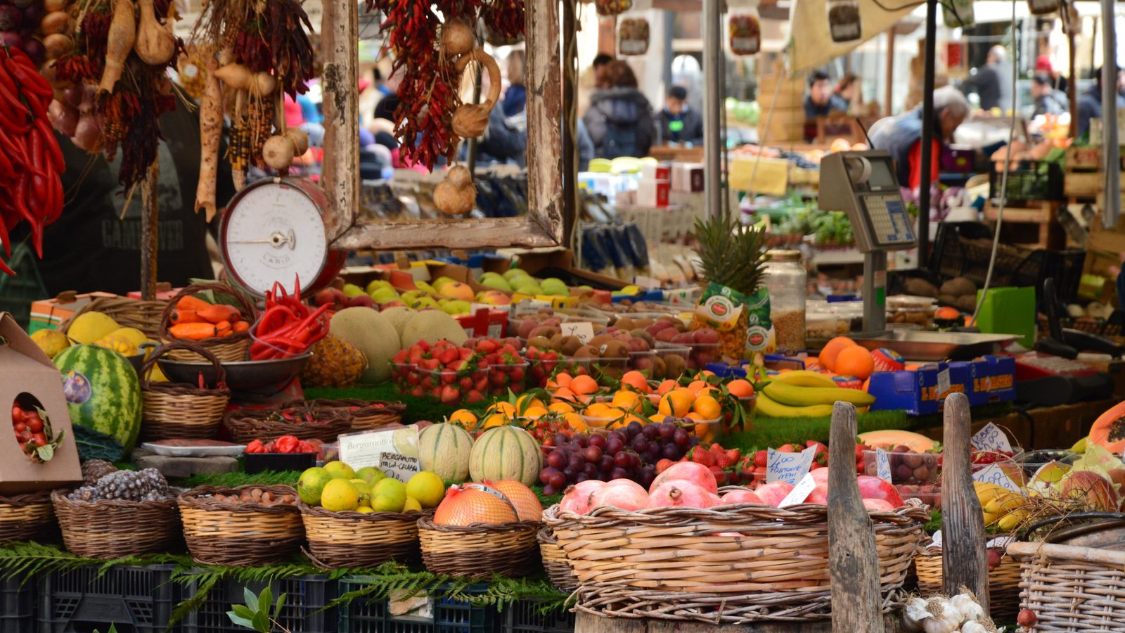 Marktstand mit Obst und Gemüse in Weidenkörben, darüber getrocknete Chilis und eine alte Waage