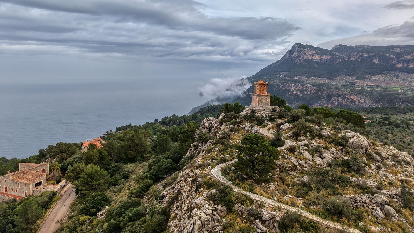 Gepflasterter Weg zu einem Aussichtsturm über der Steilküste von Mallorca, dahinter Berge und Wolken